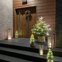 Oświetlona, drewniana weranda z trzema schodami, udekorowana na święta: drewniane drzwi z wieńcem, choinki w jutowych workach i lampiony ze świecami.