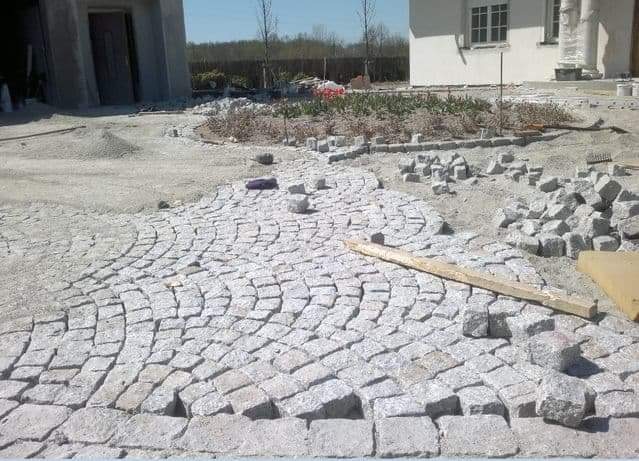 Fragment podjazdu z jasnoszarej kostki granitowej ułożonej w łuki, widoczne przygotowane kamienie, w tle budynek w stanie surowym i fragment domu.