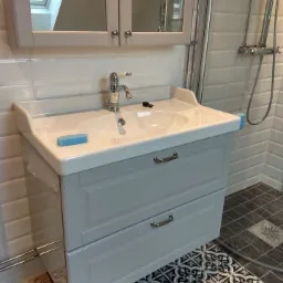 Szara szafka pod umywalkę z dwoma szufladami, chromowana bateria, białe płytki na ścianie i wzorzysta podłoga w łazience z kabiną prysznicową.