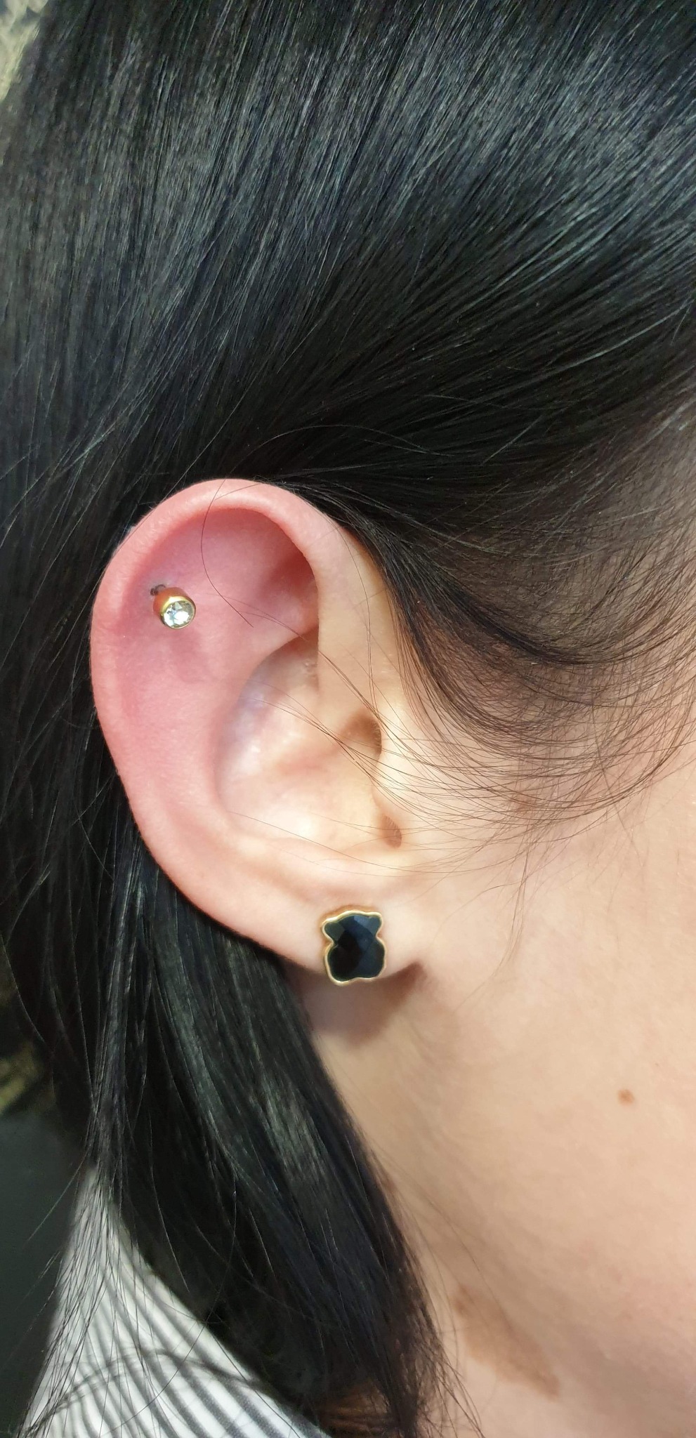 Ucho z nowym kolczykiem typu helix z małym kamieniem szlachetnym oraz czarną kolczykiem w kształcie misia w płatku ucha, widoczne ciemne włosy.