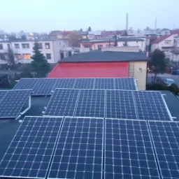 Instalacja paneli fotowoltaicznych na płaskim dachu domu jednorodzinnego w pochmurny dzień, widok na okoliczne budynki i ulicę z zaparkowanymi samochodami.