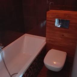 Prb Fakmet - Nowoczesna łazienka z wiszącą toaletą obudowaną drewnianą konstrukcją, mozaiką zdobiona obudowa wanny i ciemne płytki na ścianach.