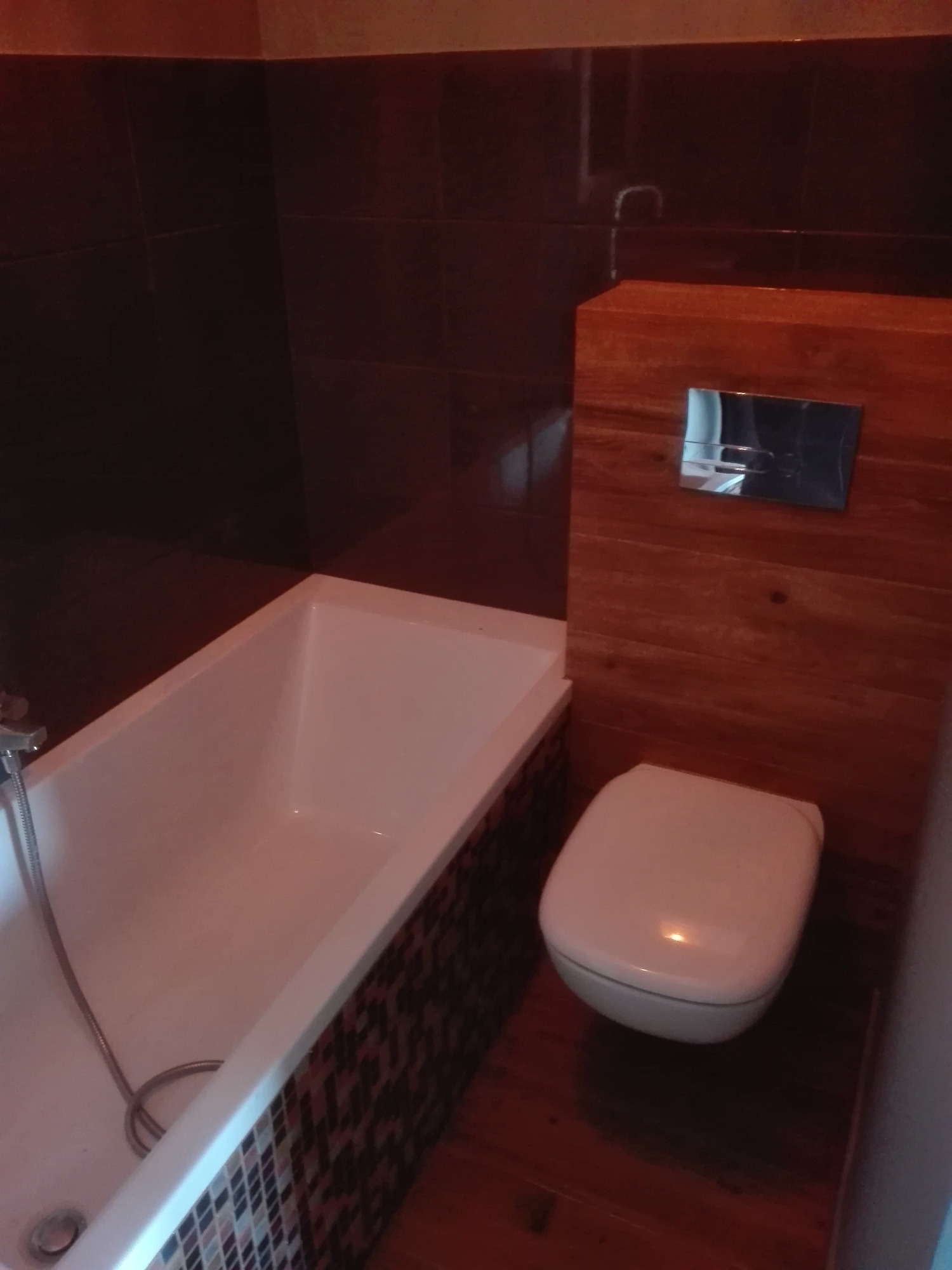 Nowoczesna łazienka z wiszącą toaletą obudowaną drewnianą konstrukcją, mozaiką zdobiona obudowa wanny i ciemne płytki na ścianach.