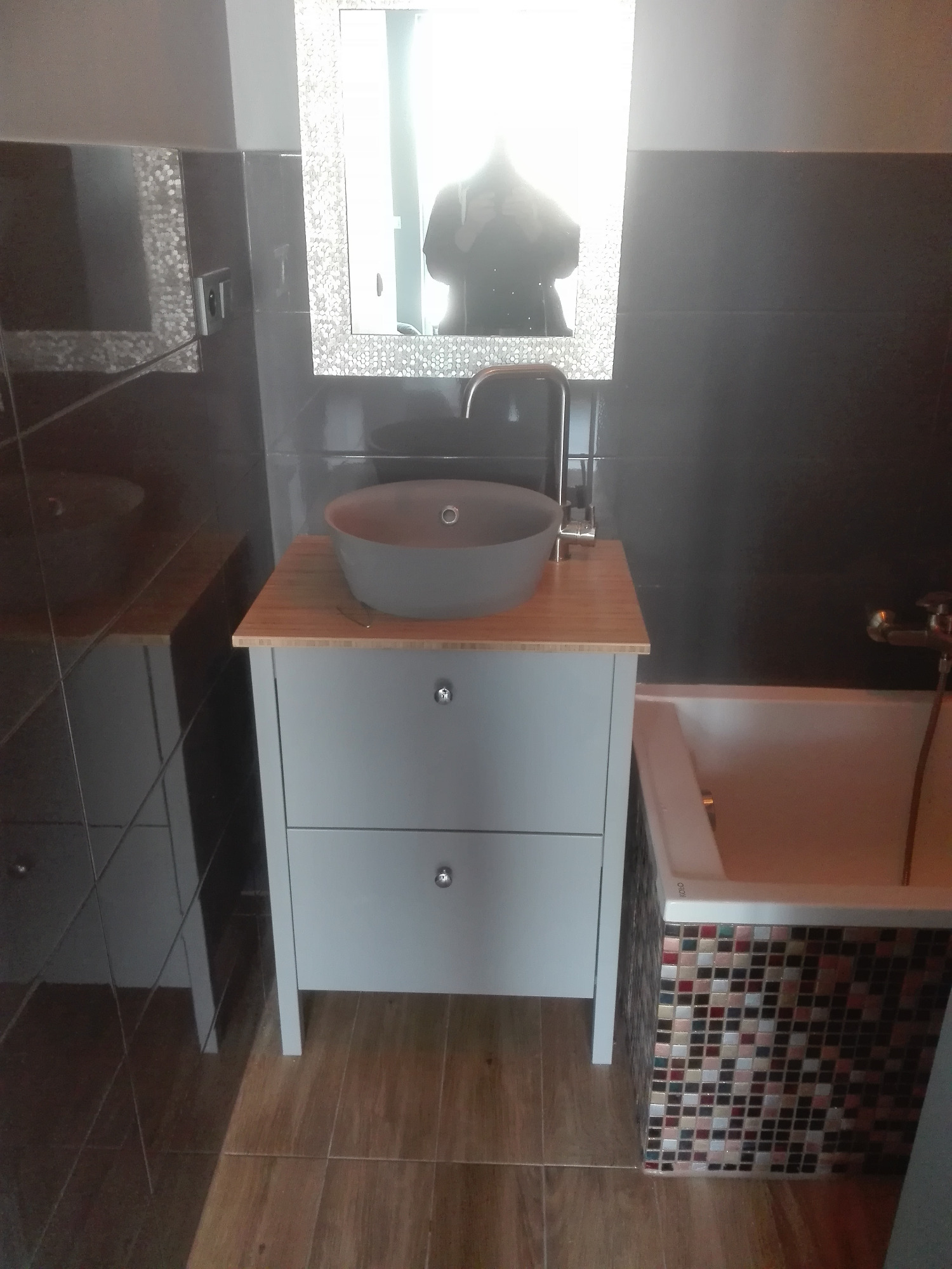 Umywalka nablatowa w kolorze szarym na szafce z dwoma szufladami z bambusowym blatem, bateria chromowana, lustro w ozdobnej ramie, ciemne płytki na ścianie, mozaika na obudowie wanny, drewnopodobne...