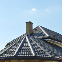 Czarny dach z dachówki ceramicznej z kominem z cegły na tle błękitnego nieba.