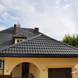 Dom jednorodzinny z ciemnoszarą dachówką, brązowymi kominami i beżową elewacją w słoneczny dzień. Widoczne rynny w kolorze dachu.