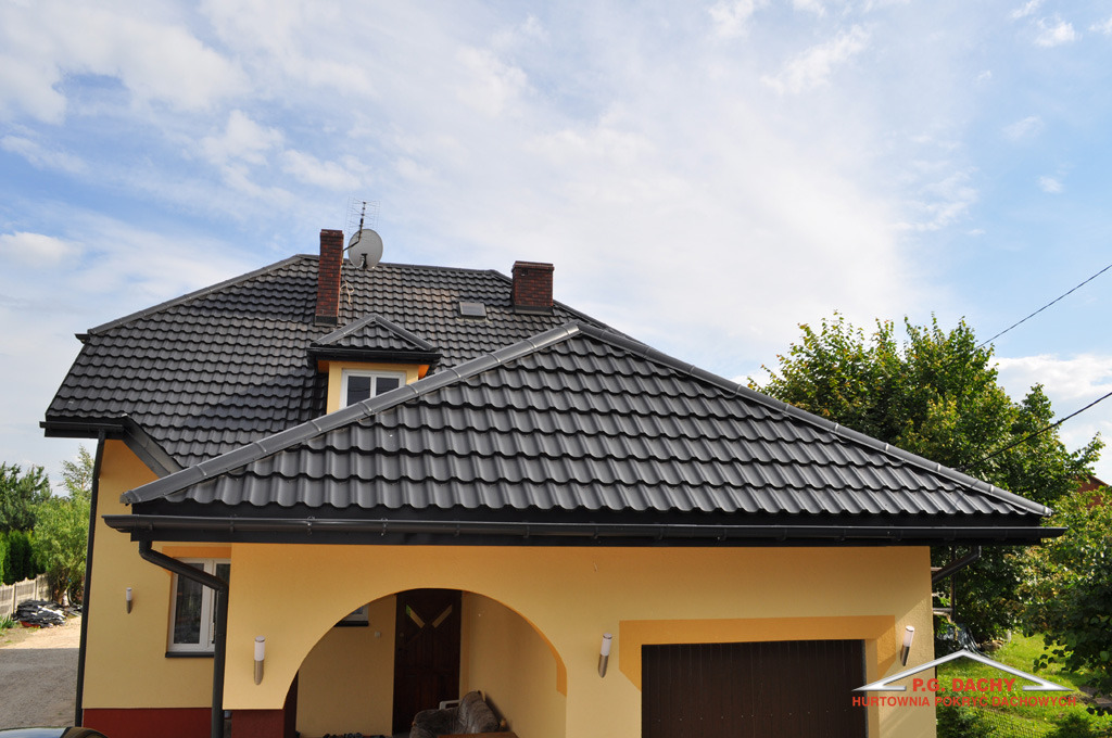 Dom jednorodzinny z ciemnoszarą dachówką, brązowymi kominami i beżową elewacją w słoneczny dzień. Widoczne rynny w kolorze dachu.