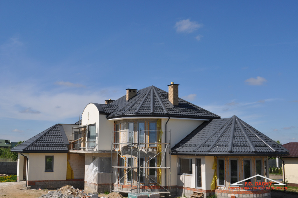 Dom jednorodzinny w trakcie budowy, z widocznym dachem pokrytym ciemnoszarą dachówką ceramiczną, kominy z cegły, elewacja w jasnym odcieniu, rusztowania przy oknach, logo firmy dekarskiej w prawym...