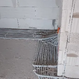 Szereg szarych, karbowanych rurek ochronnych kabli elektrycznych ułożonych równolegle na surowym, betonowym podłożu wzdłuż ściany z białych bloczków, widoczne pomarańczowe puszki instalacyjne...