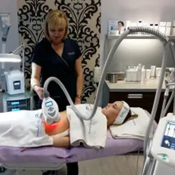 Klientka leży na ł&oacute;żku zabiegowym podczas sesji z urządzeniem do modelowania sylwetki; widoczna głowica urządzenia emitująca czerwone światło, obsługiwana przez kosmetologa w ciemnym uniformie.