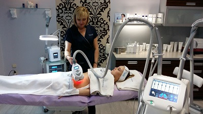 Klientka leży na łóżku zabiegowym podczas sesji z urządzeniem do modelowania sylwetki; widoczna głowica urządzenia emitująca czerwone światło, obsługiwana przez kosmetologa w ciemnym uniformie.