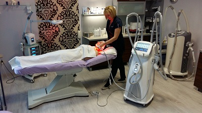 Klientka leżąca na łóżku kosmetycznym poddawana zabiegowi z użyciem urządzenia emitującego czerwone światło; w tle widoczne inne specjalistyczne urządzenia i szafki z kosmetykami w gabinecie.