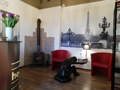 Wnętrze salonu kosmetycznego z recepcją, piecem wolnostojącym, dwoma czerwonymi fotelami, rzeźbą kobiety i fototapetą z wieżą Eiffla.
