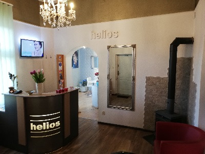 Wnętrze salonu z recepcją z logo 'helios', lustrem w srebrnej ramie, czarnym piecem kaflowym i widokiem na dalszą część salonu przez łukowate przejście.