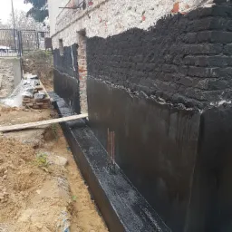 Fundament budynku z nałożoną, czarną izolacją przeciwwilgociową; widoczne wystające pręty zbrojeniowe i wykop z ziemią.
