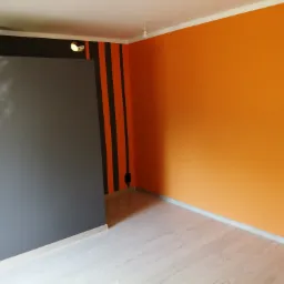 Pokój po remoncie: pomarańczowa ściana, szare pionowe pasy na ścianie obok szarej szafy wnękowej, jasna podłoga, widoczna lampa na suficie.