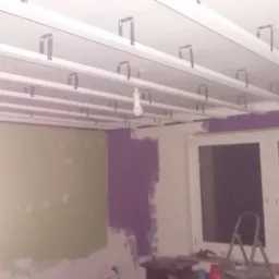 Montaż metalowej konstrukcji sufitu podwieszanego w pokoju z częściowo pomalowanymi ścianami na zielono i fioletowo, widoczna żarówka wisząca na kablu.