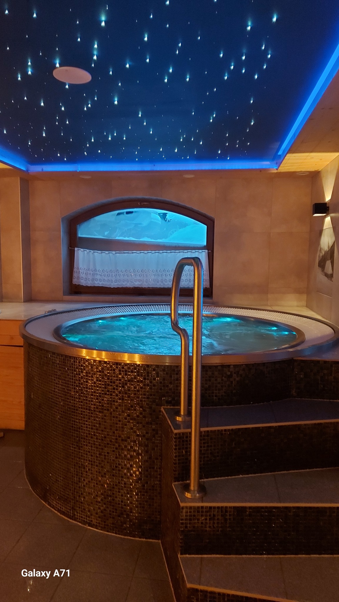 Okrągłe jacuzzi z mozaiką, oświetlone od dołu, z metalową poręczą i schodkami, pod sufitem imitującym rozgwieżdżone niebo z niebieskim podświetleniem, widok przez okno na zaśnieżony krajobraz.