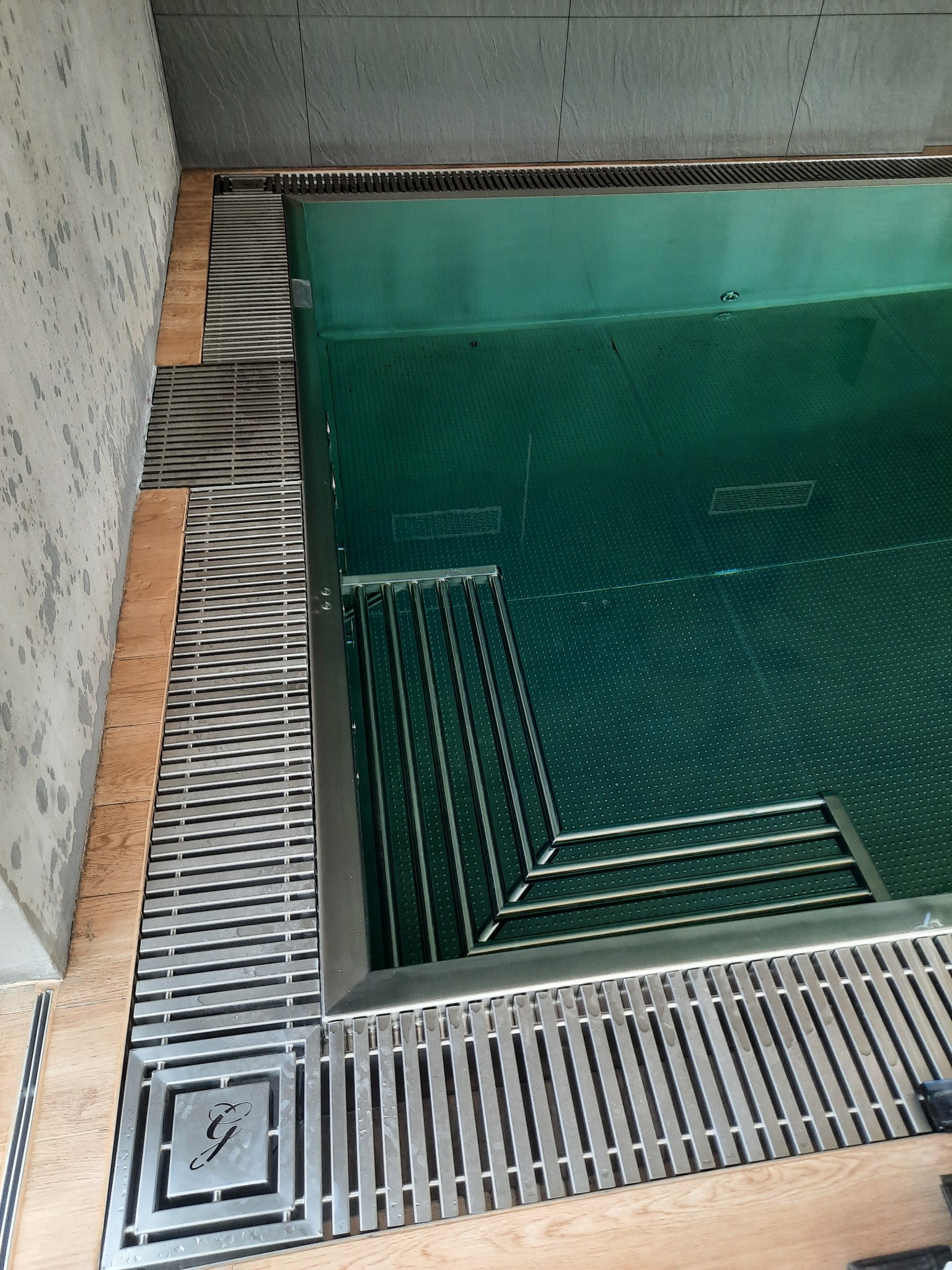 Wnętrze basenu z zieloną wodą, metalowe schodki wejściowe, kratki odpływowe z logo w rogu.