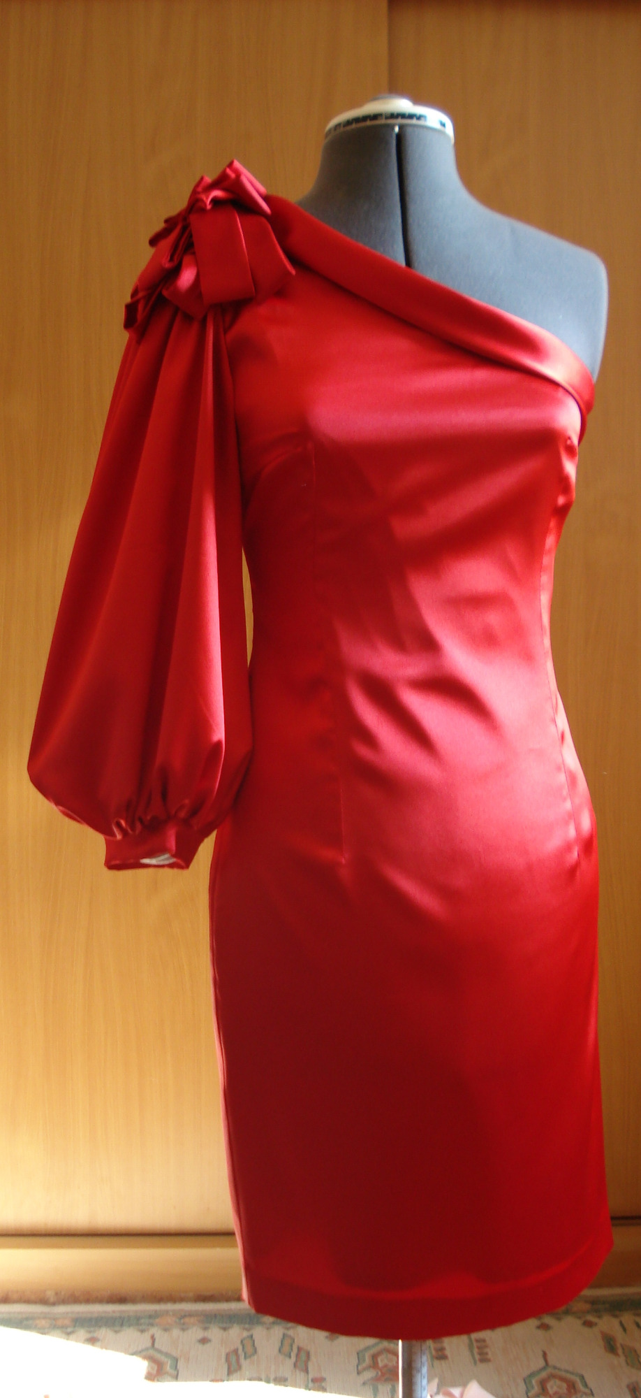 Czerwona, satynowa sukienka na jedno ramię z dekoracyjnym kwiatem, prezentowana na manekinie w pracowni w Oławie. Widoczny etap szycia lub dopasowywania odzieży.