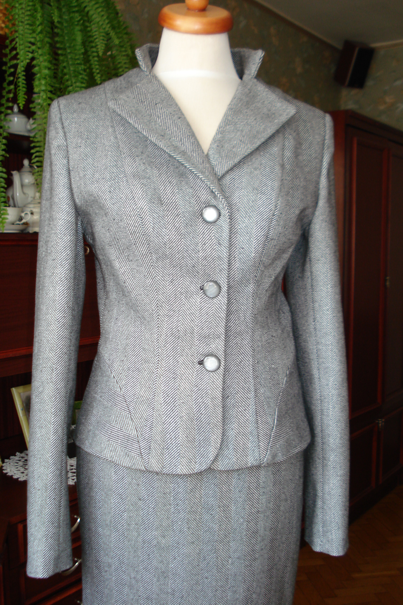 Szary damski kostium w jodełkę, składający się z marynarki zapinanej na trzy guziki i spódnicy, prezentowany na manekinie na tle mebli.