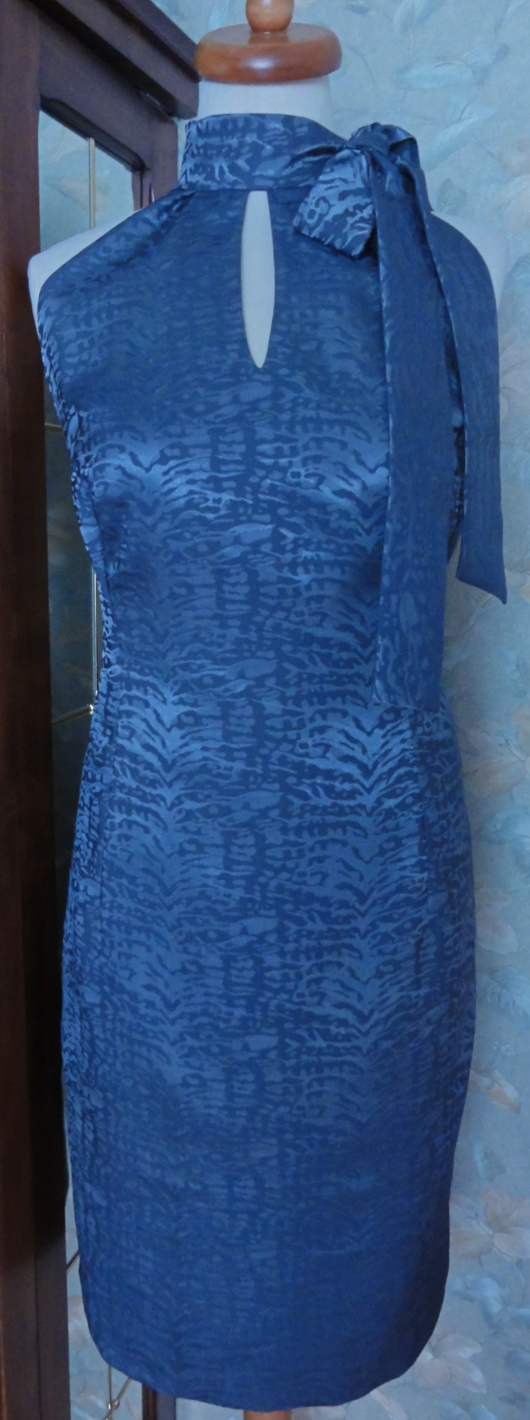 Dopasowana, niebieska sukienka z wysokim dekoltem, rozcięciem i ozdobną kokardą, prezentowana na manekinie.