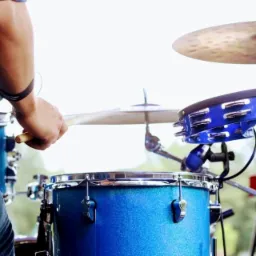 Perkusista w niebieskiej koszuli w kratę uderza pałeczką w talerz perkusyjny, widoczny niebieski bęben z brokatem i tamburyn.