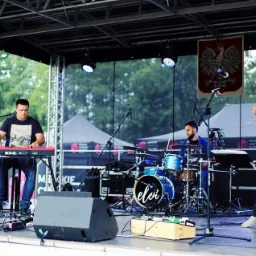 Zespół muzyczny Eloi podczas występu na scenie plenerowej, pianista gra na keyboardzie Nord Electro 5D, perkusista za bębnami, gitarzysta w czerwonej koszuli w kratę gra na gitarze akustycznej,...