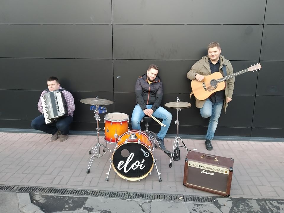 Trzech muzyków pozuje z instrumentami: akordeon, perkusja z logo 'eloi' i gitara akustyczna, obok wzmacniacz Marshall, na tle ciemnej ściany.