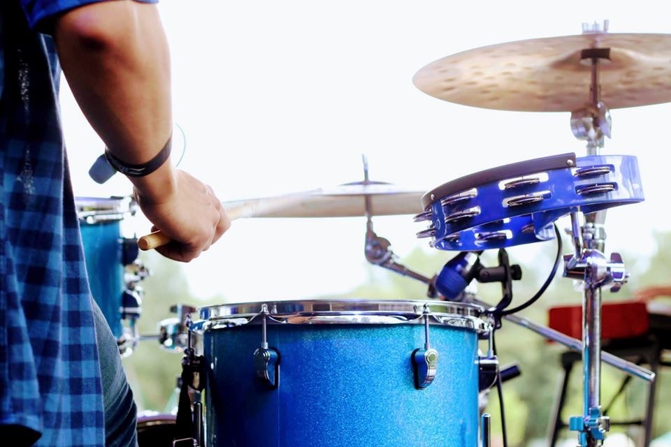 Perkusista w niebieskiej koszuli w kratę uderza pałeczką w talerz perkusyjny, widoczny niebieski bęben z brokatem i tamburyn.