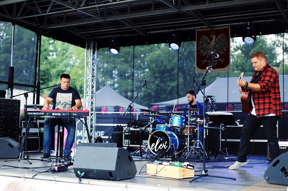 Zespół muzyczny Eloi podczas występu na scenie plenerowej, pianista gra na keyboardzie Nord Electro 5D, perkusista za bębnami, gitarzysta w czerwonej koszuli w kratę gra na gitarze akustycznej,...