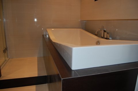 Nowoczesna, prostokątna wanna z chromowaną baterią w minimalistycznej łazience z beżowymi płytkami i ciemnobrązową obudową wanny.