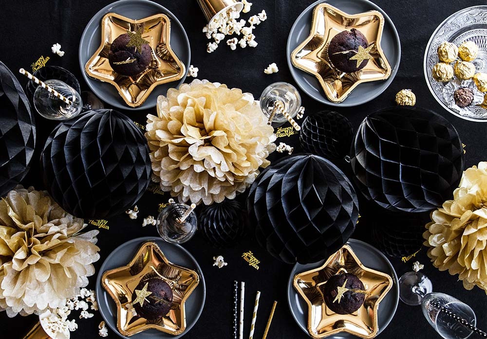 Dekoracje sylwestrowe: złote talerze w kształcie gwiazd z babeczkami, czarne i złote kule dekoracyjne, popcorn i słomki na czarnym tle.