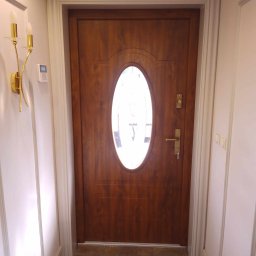 Remofleks Mateusz Ranachowski - Drewniane drzwi wejściowe z owalnym przeszkleniem, widoczne od wewnątrz, z ozdobną ościeżnicą i złotym kinkietem na ścianie.
