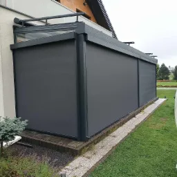 Zadaszenie tarasu z szarymi, rozwijanymi pionowymi przesłonami przeciwsłonecznymi, widoczne z zewnątrz na tle trawnika i roślinności.