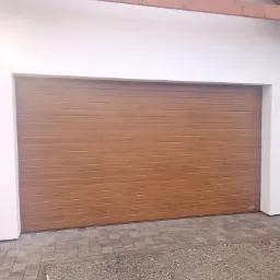 Zewnętrzna brama garażowa segmentowa w odcieniach brązu, z poziomymi przetłoczeniami imitującymi drewno, zamontowana w białej elewacji budynku z widoczną drewnianą belką stropową i brukowanym...