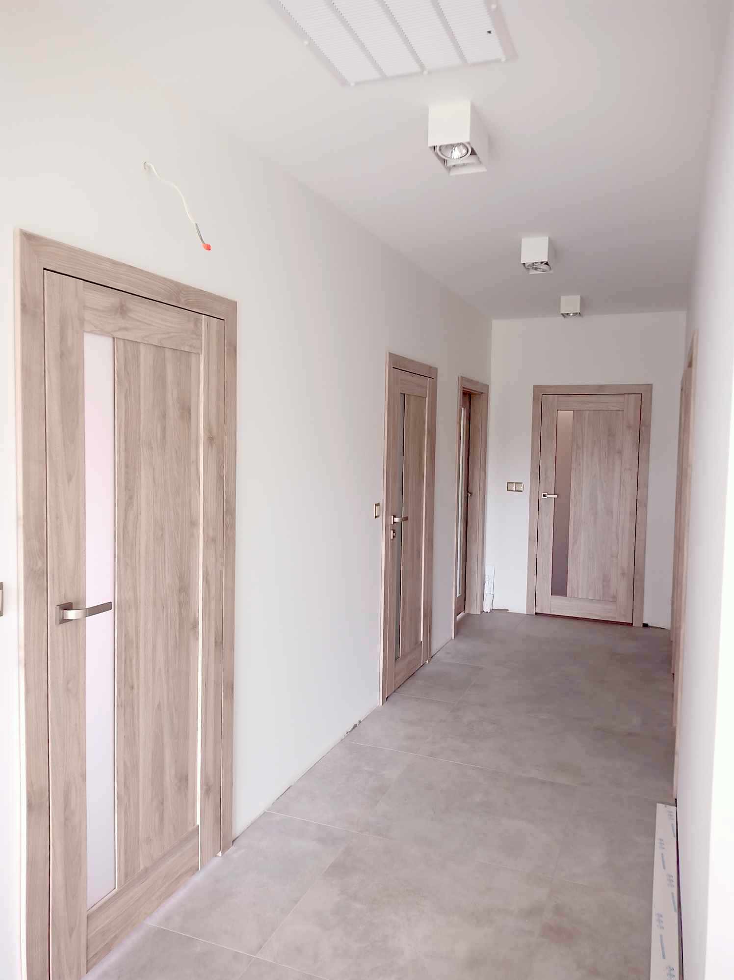 Jasne, drewnopodobne drzwi wewnętrzne z przeszkleniem w korytarzu, widoczne niedokończone prace wykończeniowe.