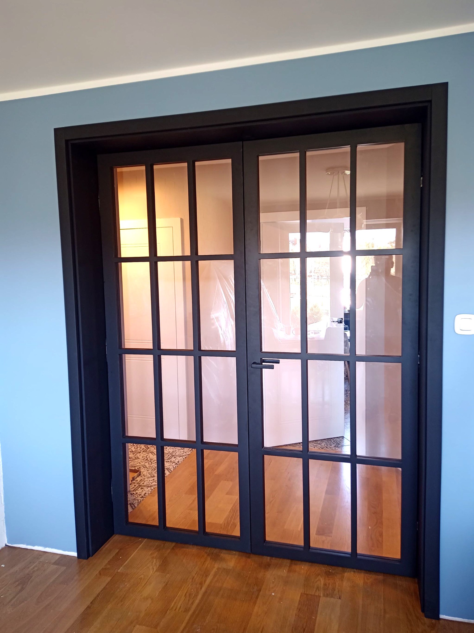 Dwuskrzydłowe, przeszklone drzwi wewnętrzne z czarną ramą i szprosami, widoczne na tle błękitnej ściany i drewnianej podłogi, z refleksami światła słonecznego na szybach.