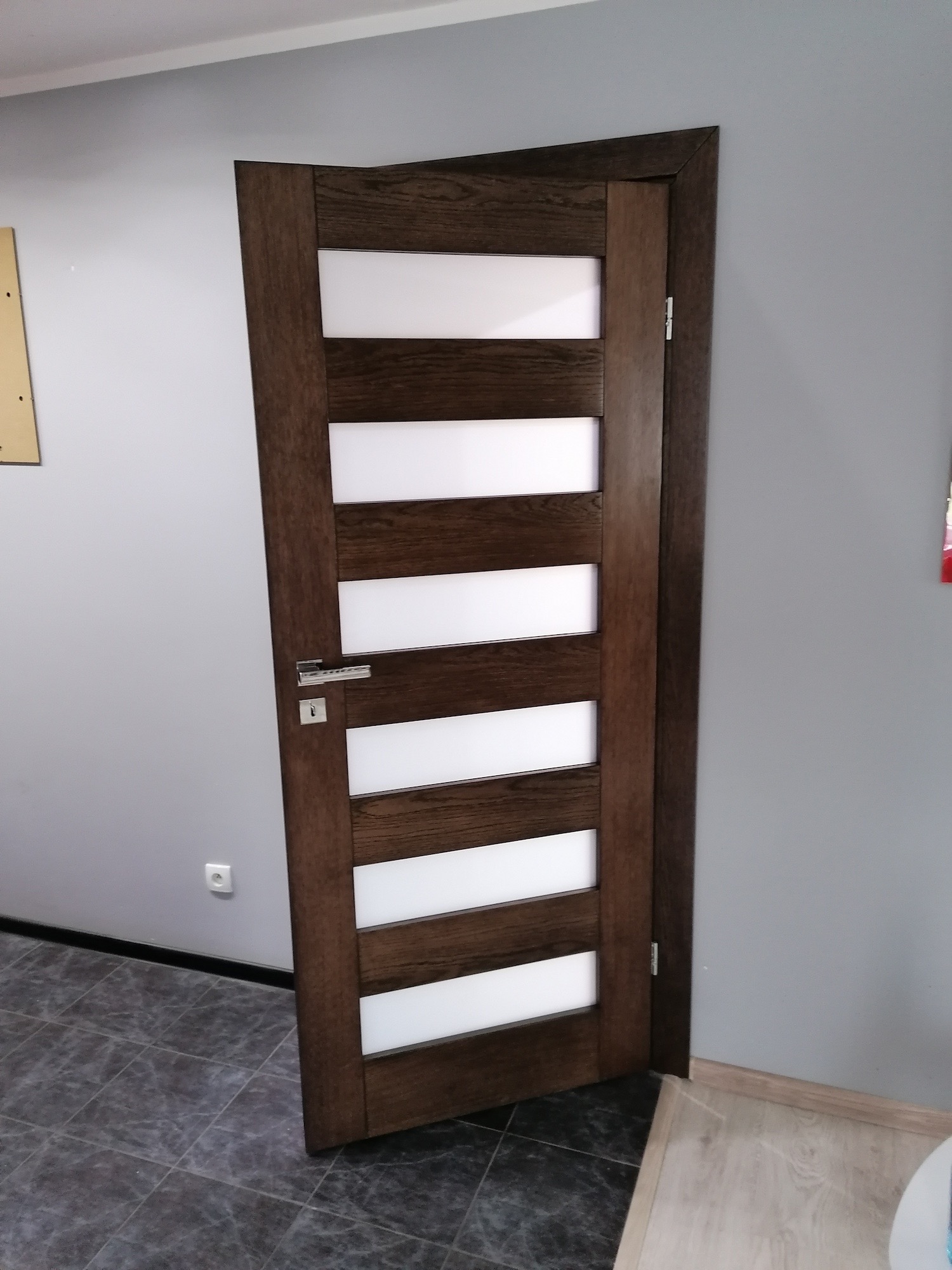 Brązowe drzwi wewnętrzne z przeszkleniami w poziomych pasach, widoczne klamka i zamek, na podłodze szara terakota imitująca kamień.