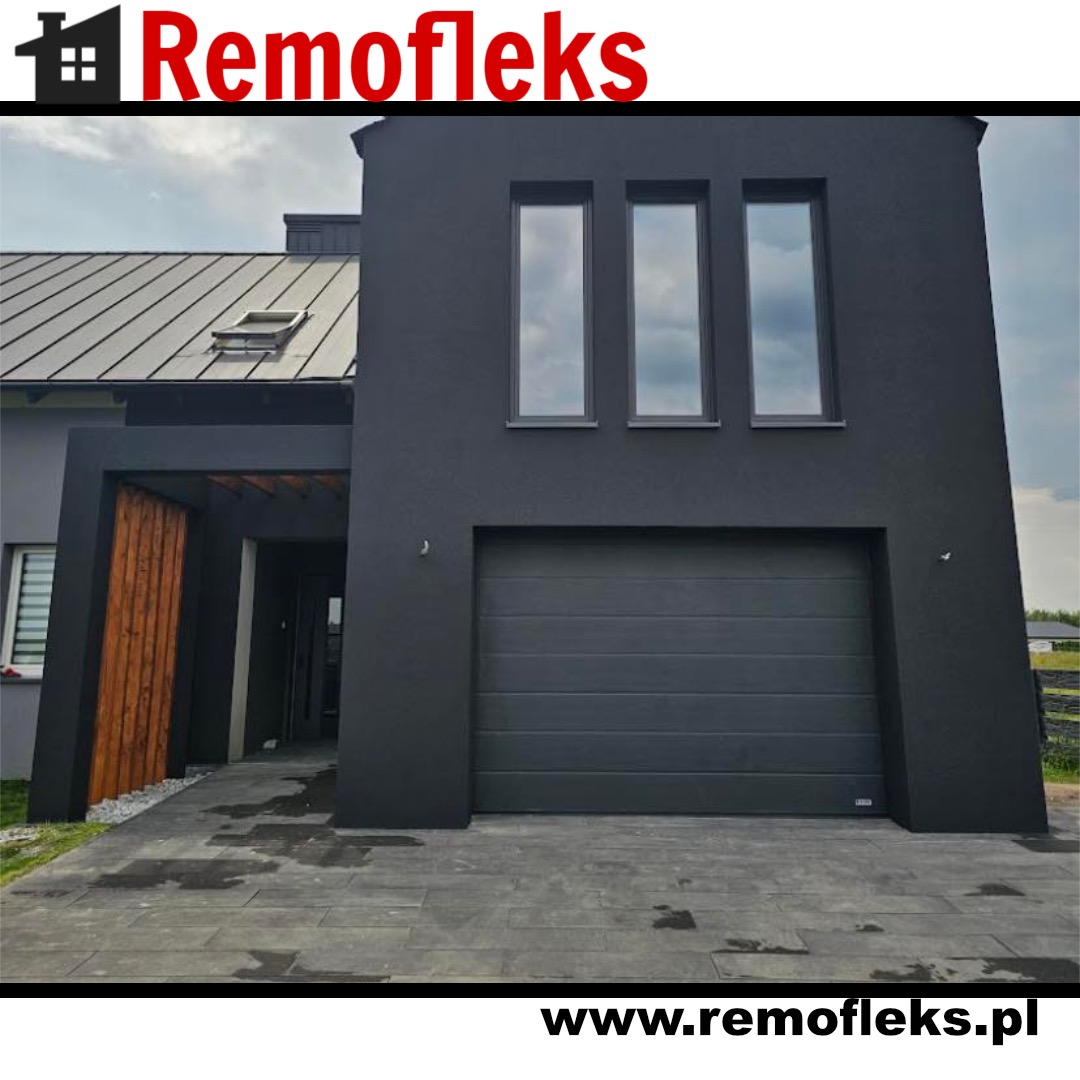 Nowoczesny dom z antracytową elewacją, trzema oknami na piętrze i segmentową bramą garażową w tym samym kolorze, widoczny fragment zadaszonego wejścia z drewnianymi elementami oraz bruk przed garażem.