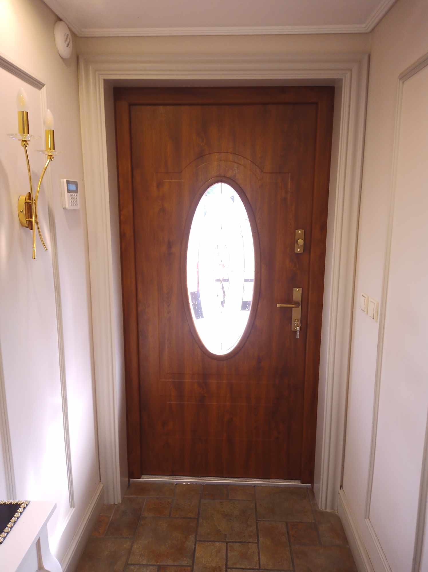 Drewniane drzwi wejściowe z owalnym przeszkleniem, widoczne od wewnątrz, z ozdobną ościeżnicą i złotym kinkietem na ścianie.