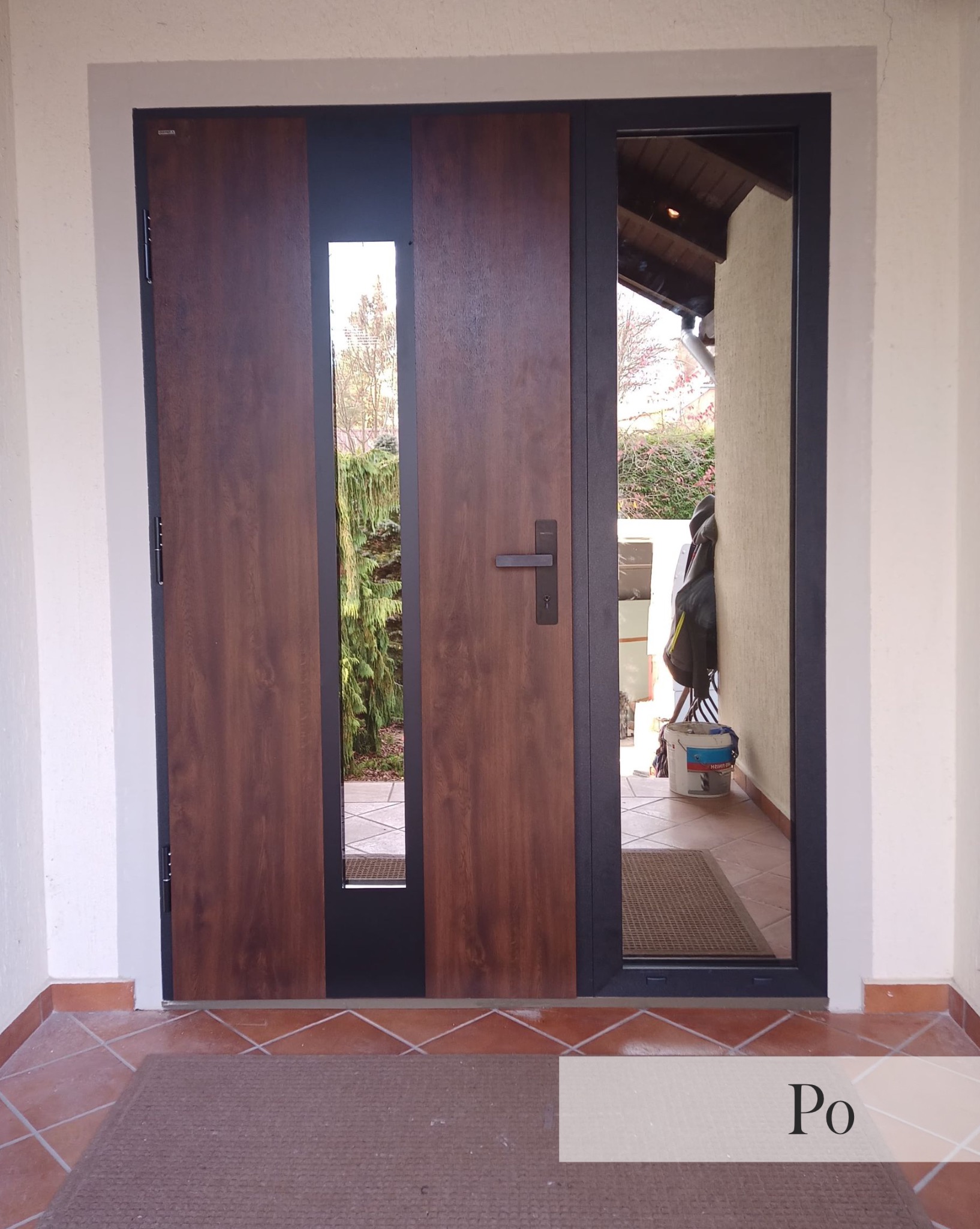 Nowoczesne drzwi wejściowe z drewnianym panelem i przeszkleniem, widok z zewnątrz na wejście do domu z widocznym fragmentem ogrodu i elementami wyposażenia.