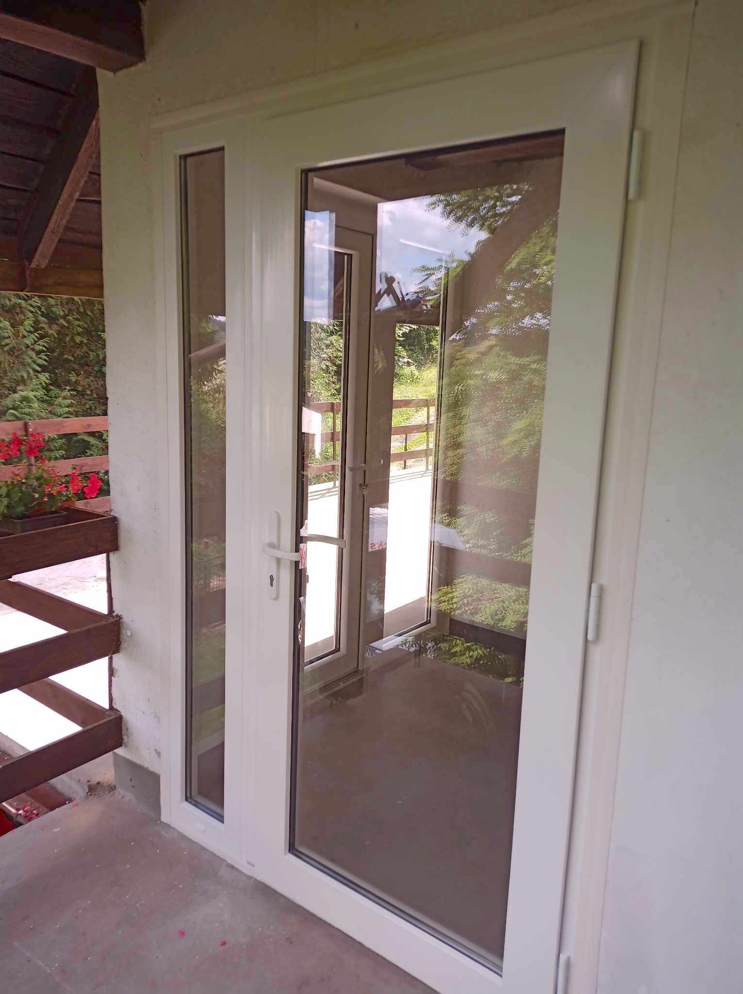 Białe drzwi balkonowe PCV z szybą zespoloną, widok z zewnątrz, z odbiciem zieleni w szybie i drewnianą balustradą z kwiatami po lewej stronie.