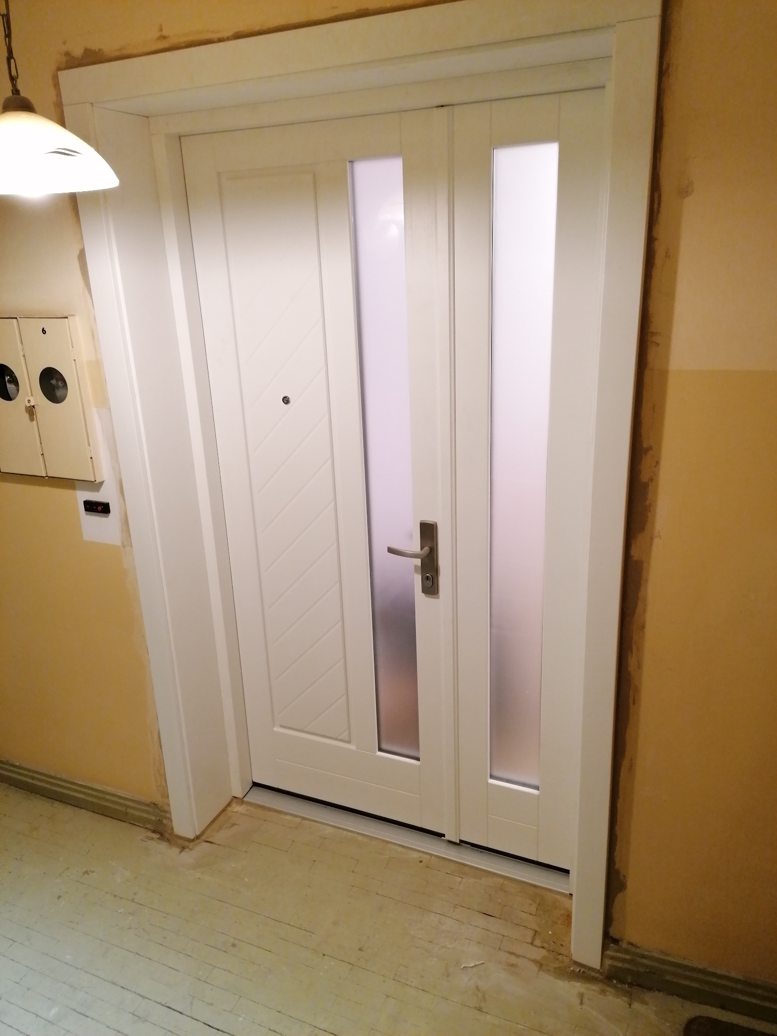 Białe drzwi wewnętrzne z dwoma pionowymi szybami z matowego szkła i ukośnym wzorem na panelu, zamontowane w odnowionym mieszkaniu z żółtymi ścianami i pomalowaną podłogą.