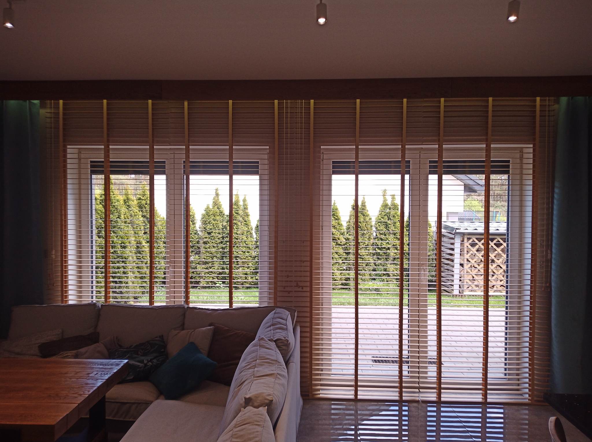 Salon z dwoma oknami przesłoniętymi drewnianymi żaluzjami poziomymi, widok na ogród z tujami i drewutnią.