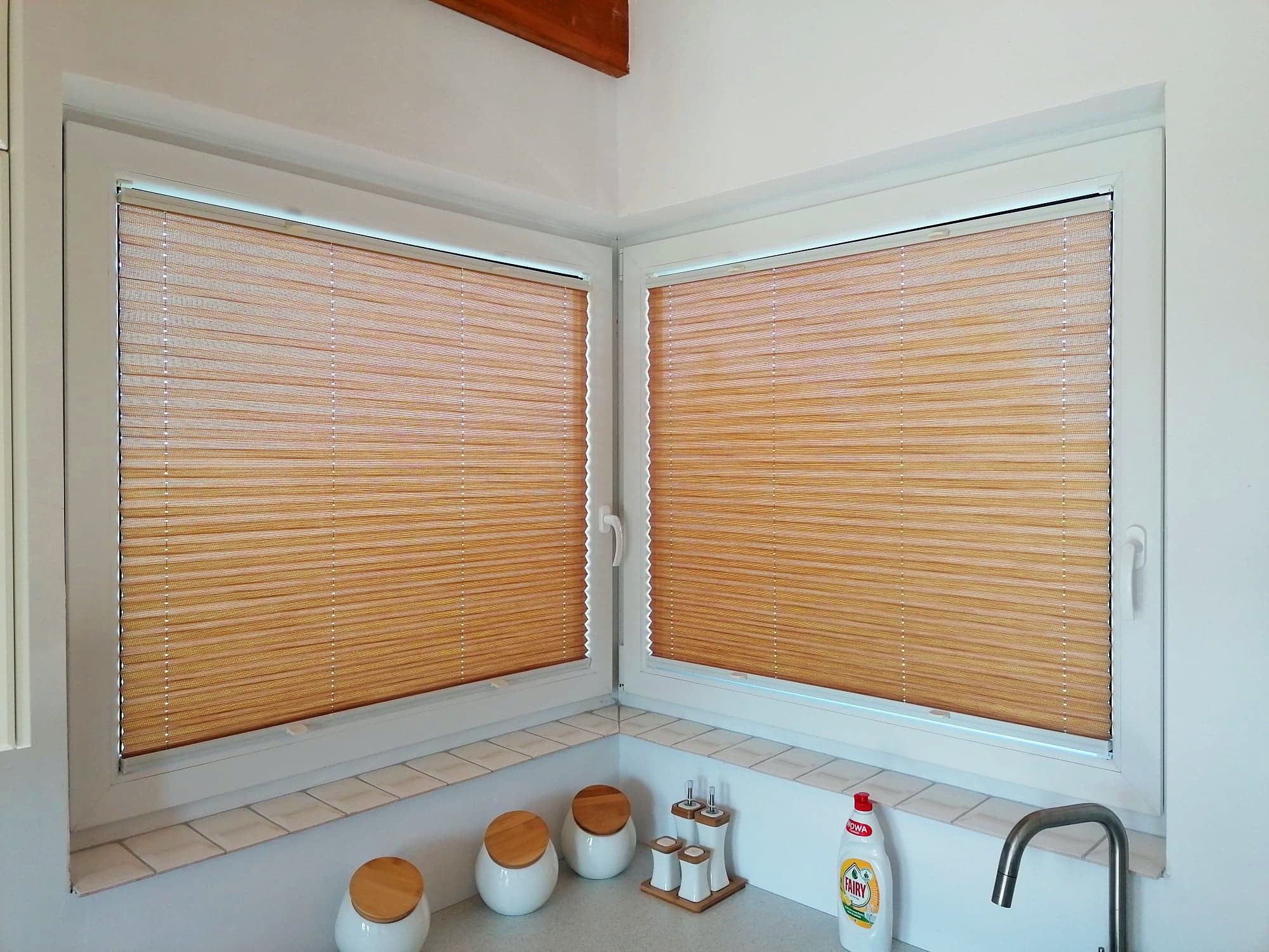 Dwa białe okna PCV narożne z zamontowanymi plisowanymi, pomarańczowymi roletami wewnętrznymi w kuchni, widoczne ceramiczne pojemniki i płyn do naczyń.