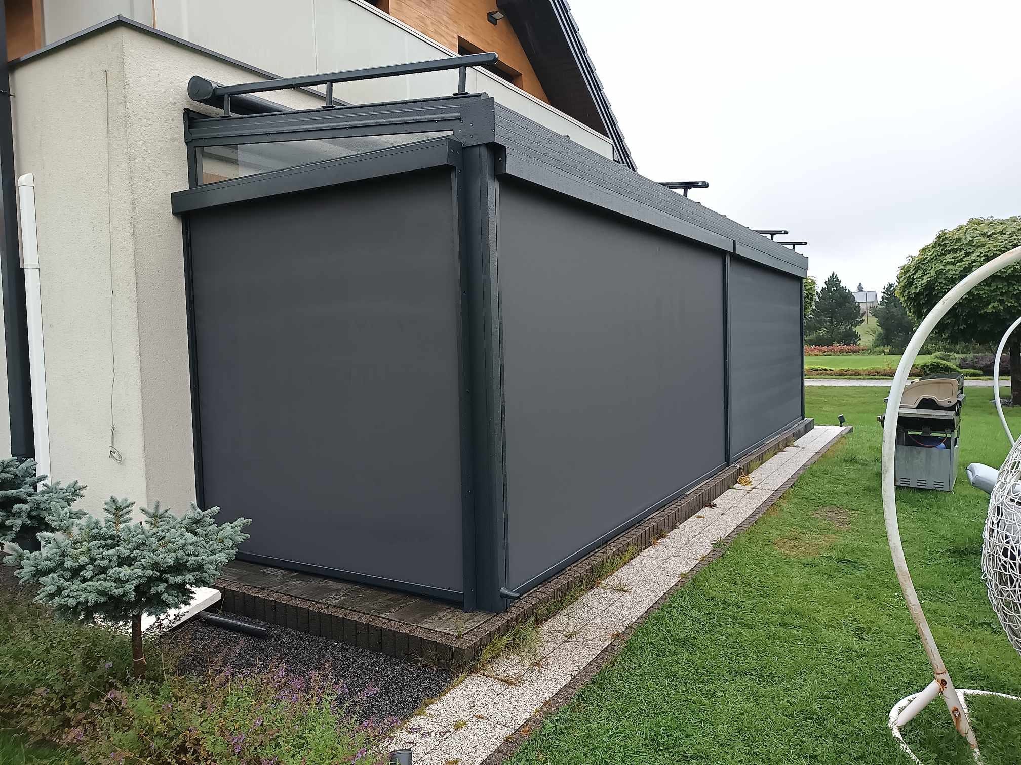 Zadaszenie tarasu z szarymi, rozwijanymi pionowymi przesłonami przeciwsłonecznymi, widoczne z zewnątrz na tle trawnika i roślinności.
