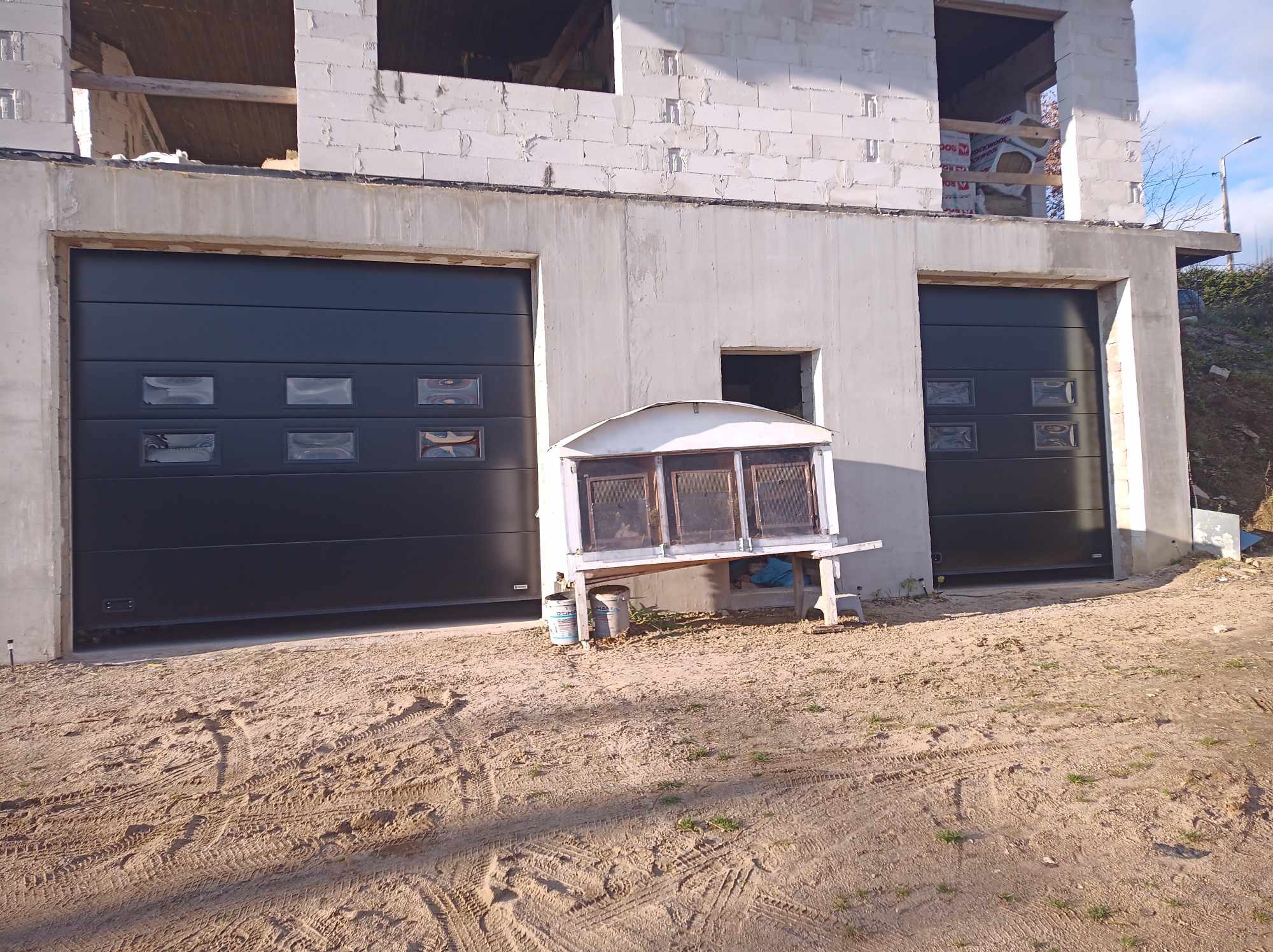 Dwie czarne bramy garażowe segmentowe z oknami w nowym, surowym budynku z betonu komórkowego, na piaszczystym podłożu widoczne ślady opon, obok stoi stary stół warsztatowy z farbami.