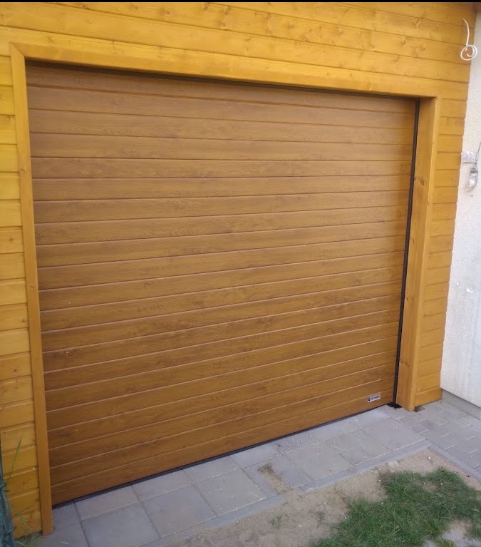 Zamknięta brama garażowa segmentowa w kolorze jasnego drewna, zamontowana w drewnianej elewacji budynku, widoczna z zewnątrz na tle szarej kostki brukowej.