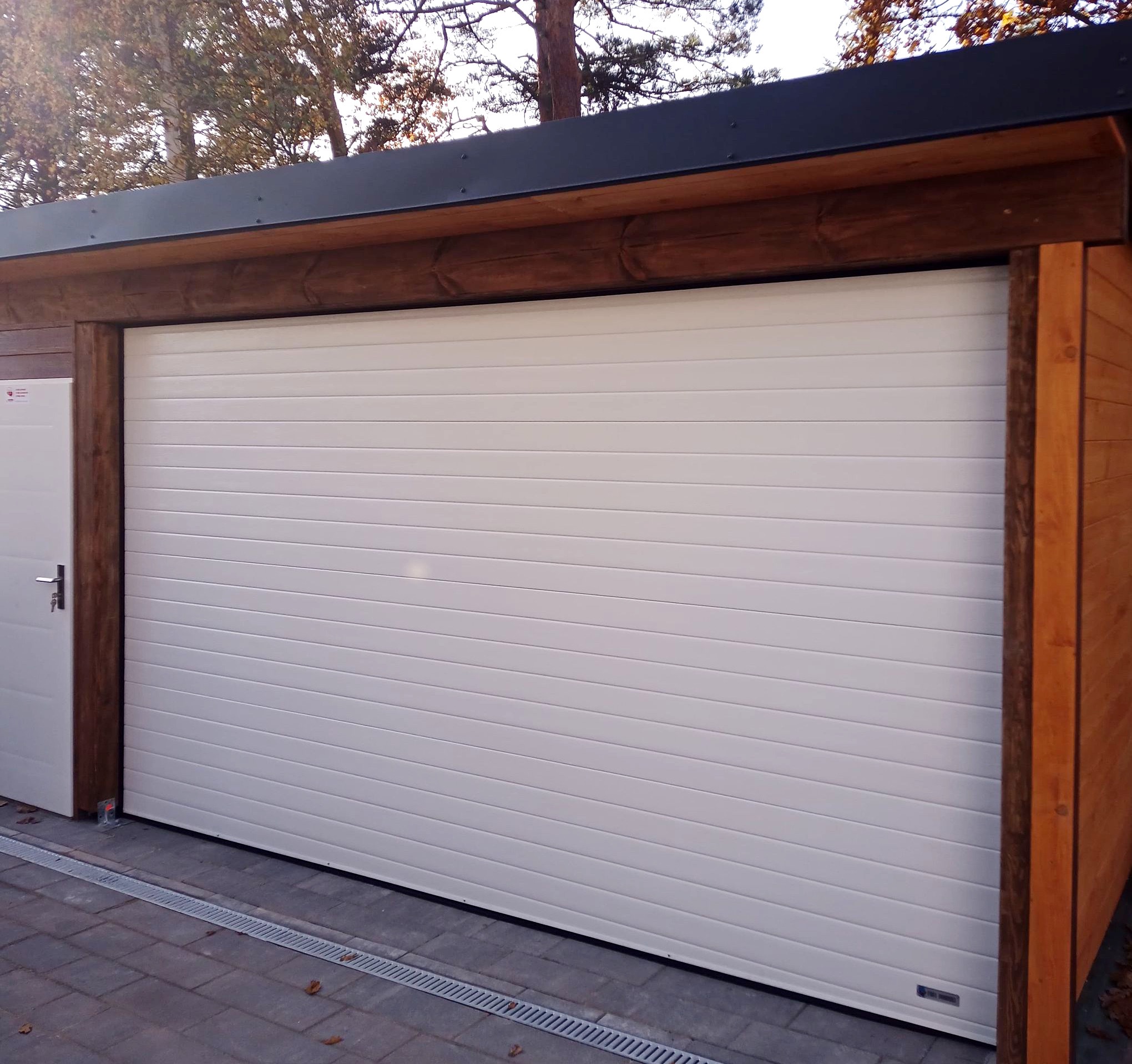 Biała segmentowa brama garażowa z drewnianą ramą, widoczna z zewnątrz, zamontowana w nowoczesnym garażu z desek.